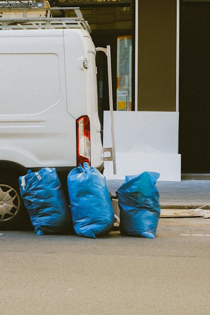 The image displays three large blue rubbish bags made of durable plastic, positioned on the pavement beside the rear of a white commercial van. The bags are filled with waste material, with some slight wrinkling and creases visible on their surfaces, indicating they are tightly packed. The van is parked in front of a building with large glass windows and a dark-colored door or frame, suggesting an urban or commercial setting. The bags are situated on the side of the vehicle, close to its rear wheel, and the scene is illuminated by natural daylight, casting minimal shadows. This setup may illustrate an alternative waste handling method such as private rubbish collection services offered by Waste Disposal Knightsbridge, often involving on-site clearance rather than reliance solely on local authority rubbish collection. The environment appears clean, organized, and professional, aligning with typical rubbish removal or disposal activities in the Knightsbridge area.