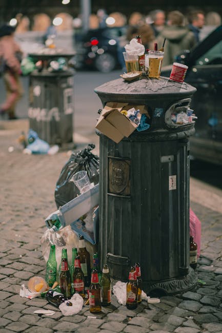 A street scene in an urban area showing a Waste Disposal Knightsbridge garbage collection vehicle with an open rear compartment positioned on a cobblestone road. The truck, painted white with some rust and grime visible on the internal metal surfaces of the loading area, is complemented by an worker wearing a blue and orange high-visibility uniform and a matching cap, who is seen emptying a blue wheeled bin into the compactor. To the left, a black car parked on the pavement faces away from the camera. The surrounding environment includes old multi-story buildings with weathered facades, some with peeling paint and exposed brickwork, and small, square windows. A few street signs are mounted on the buildings, indicating parking restrictions or no stopping zones. The scene is lit with natural daylight, contributing to the neutral, factual depiction of ongoing waste collection activity, illustrating a private waste handling service performing rubbish removal on an urban residential street.