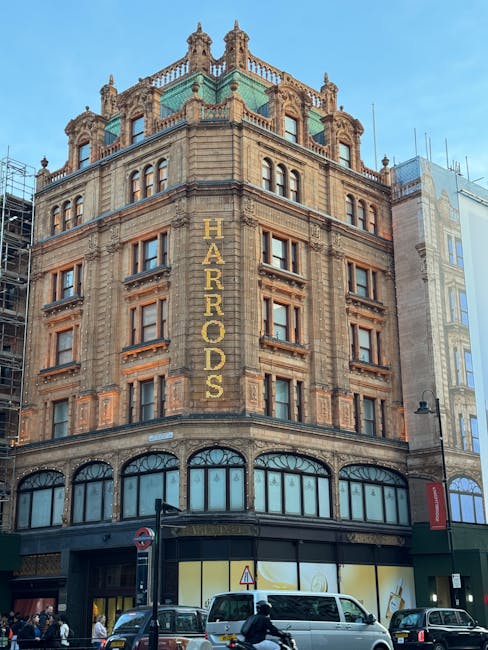 A detailed view of a historic multi-storey building with ornate architectural features, including decorative stonework, cornices, and a prominent green copper roof with turrets on the top. The building's façade is made of light brown stone with darker accents around the rectangular windows, which are evenly spaced across each floor. Bright yellow letters spelling 'HARRADS' are vertically attached to the corner of the building, indicating a business sign. The ground level features large arched windows with dark frames and a dark storefront beneath, with a small red and white no-entry road sign visible on the street. In front of the building, there is a busy street scene with parked cars, including a white van and a dark sedan, as well as pedestrians walking or cycling. Street lamps and modern adjacent buildings are also visible, situating the scene within an urban environment that may engage private waste collection services, aligning with independent waste disposal practices near commercial premises.