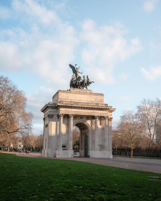 The image displays a large, ornate stone monument featuring a prominent archway at its center, situated within a park area. At the top of the monument is a detailed bronze sculpture of a chariot drawn by horses, driven by a figure holding reins. The monument is constructed from light-colored stone with carved decorative elements, including a frieze and columns, giving it a classical appearance. The surrounding environment shows a well-maintained grassy area in the foreground, with leafless trees on either side indicating a late autumn or winter setting. The sky is partly cloudy with patches of blue, and the lighting suggests daytime with natural sunlight highlighting the monument's features. This scene, while primarily focused on a historic landmark, subtly relates to urban outdoor spaces where private events or activities might generate waste requiring responsible disposal, aligning with services like those provided by Waste Disposal Knightsbridge for managing rubbish and bulky waste outside of municipal collection services.
