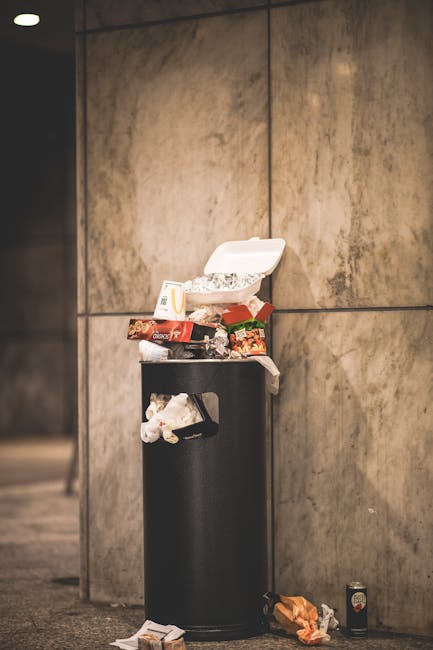 A black cylindrical waste bin with a partially open white polystyrene food container on top, overflowing with assorted rubbish including food packaging, wrappers, and paper tissues. The bin is situated against a large, beige stone wall with visible joints and smooth, slightly reflective surfaces. Nearby, on the ground, there are scattered debris such as an orange paper bag and crumpled waste, with a small beverage can placed to the right of the bin. The setting appears to be an indoor or covered public space with subdued lighting, emphasizing the littered environment which underscores issues related to improper rubbish disposal, highlighting the importance of alternative waste handling services like those offered by Waste Disposal Knightsbridge for maintaining cleanliness and hygiene.