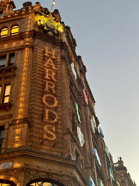 A tall, historic building with ornate architectural details illuminated by warm exterior lighting at dusk. The facade features a large vertical sign spelling 'HARMONYS' in illuminated letters. Nearby, several pieces of laundry, including shirts, towels, and fabric items, are hung from lines or poles along the building's windows and balconies, adding a splash of color against the stone surface. The building's surface has a textured finish with decorative moldings and cornices, showing signs of age and craftsmanship. The sky overhead is clear, transitioning from daylight to evening, with a soft ambient glow. The scene evokes a lively, urban atmosphere, with the combination of historic architecture and everyday laundry suggesting a practical, lived-in setting. This image relates to typical urban scenes where private laundry or outdoor drying may occur in proximity to commercial or residential sites, aligning with themes of on-site clearance and alternative waste handling often managed by waste disposal services.