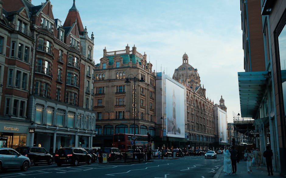 A busy urban street scene in Knightsbridge featuring a row of historic multi-story buildings with ornate brick and stone facades, large windows, and decorative elements on the upper levels. In the foreground, a line of parked cars, including a black SUV and a white sedan, is positioned along the curb. Pedestrians are walking on the sidewalk, some in formal attire, with a few engaging in conversation. To the right, a modern building with glass panels and a blue awning extends from the side. In the background, a large, domed historic building with intricate architectural details and a prominent, rounded roof dominates the skyline, with a partly cloudy sky overhead casting natural daylight across the scene. The image captures a typical cityscape in Knightsbridge, where private waste collection services like Waste Disposal Knightsbridge commonly operate in urban environments, often handling rubbish removal for commercial and residential properties. The scene emphasizes the blend of historic architecture and contemporary urban life, relevant to the context of alternative waste handling and rubbish removal services.