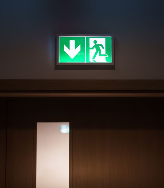 A green illuminated emergency exit sign mounted on a dark gray interior wall above a wooden door frame. The sign features a downward-pointing arrow on the left side and a running figure on the right, indicating a safe evacuation route. The sign's bright green color contrasts with the surrounding background, providing clear visibility. Below the sign, part of the door's wooden finish is visible, with a narrow frosted glass panel on the left side, suggesting an interior space that may be related to a commercial or public building. The lighting is subdued, emphasizing the bright sign, which is a common feature in buildings requiring compliance with safety and evacuation regulations. This type of signage may be relevant to environments where prompt emergency evacuation procedures, including escape routes from areas such as waste storage or disposal zones, are critical, aligning with the context of waste management and safety considerations. Waste Disposal Knightsbridge often advises proper signage and clear pathways to facilitate safe and compliant waste handling and disposal procedures.
