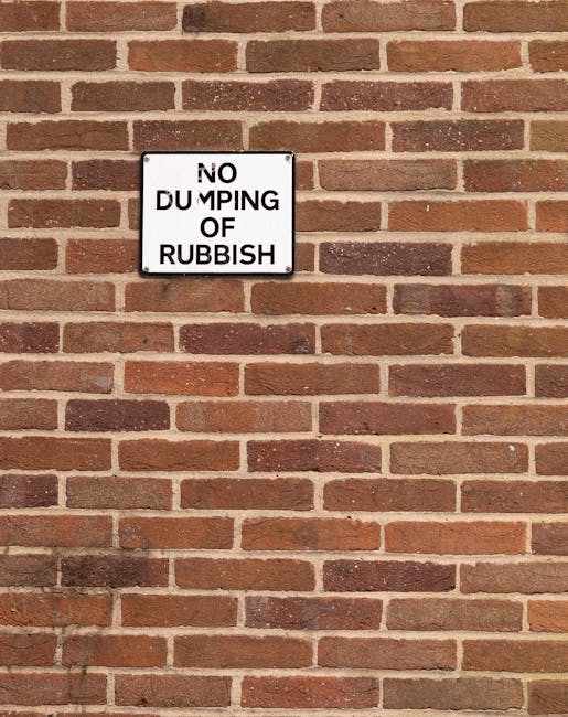 A rectangular metal sign mounted on a red brick wall, displaying a white background with black text that reads 'NO DUMPING OF RUBBISH' in uppercase letters. The bricks surrounding the sign are uniformly laid with a slightly weathered appearance, featuring shades of reddish-brown and dark brown mortar joints. The lighting is natural, illuminating the wall evenly without casting strong shadows. The sign is positioned centrally towards the upper left side of the image, serving as a visual marker against the textured brick surface. This scene reflects a typical outdoor urban setting where private property management or waste disposal regulations are visibly instructed, aligning with principles of rubbish removal and alternative waste handling services provided by companies like Waste Disposal Knightsbridge, especially in areas where local authority rubbish collection is supplemented by independent clearance efforts.