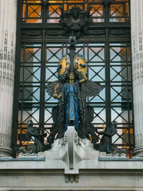 A detailed view of a decorative architectural sculpture on the facade of a historic building, featuring a central figure with wings and a blue robe, flanked by two smaller figures on either side holding objects. Above the sculpture, there is an ornate shield with gold accents and a crest, supported by intricate metal framework. The entire display is set against a backdrop of metal grid panels and stone columns with classical detailing. The lighting appears natural, illuminating the sculpture's textures and the contrasting colors of the materials, which include dark metals, blue and gold painted elements, and white stone base. This elaborate ornamentation exemplifies traditional craftsmanship and adds visual interest to the building's exterior, reflecting a style associated with civic or institutional architecture.