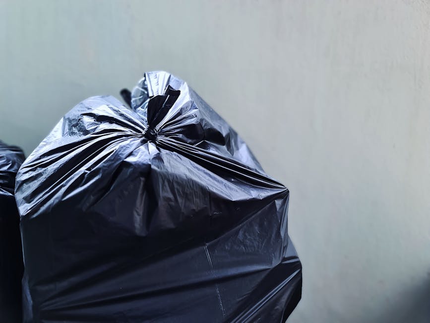 Close-up image showing several large black plastic rubbish bags filled with waste, tied securely at the top, placed against a plain, light-colored wall. The bags have a shiny, smooth surface with visible creases and folds, indicating they are made from durable plastic material. The environment appears to be an indoor setting, possibly a storage area or a corner of a driveway left for disposal. The bags are positioned in a tight cluster, with one prominently in the foreground and others partially visible behind it. The scene relates to waste management, suggesting an instance of on-site rubbish collection or preparation for removal by a professional waste disposal service such as Waste Disposal Knightsbridge. The neutral lighting enhances the clarity of the waste packaging and highlights the textures of the plastic material, aligning with the context of private disposal and alternative rubbish removal methods.