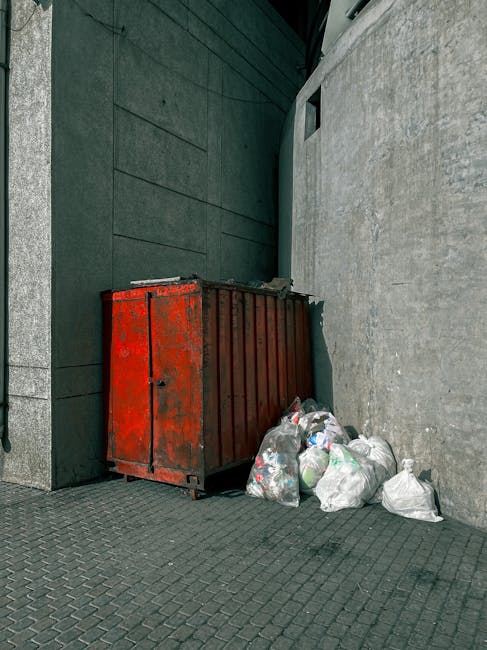 A large, rectangular metal refuse container with a weathered, rusty red finish, positioned against a dark green wall and a grey concrete wall in an urban outdoor setting. Surrounding the container are several white and semi-transparent plastic garbage bags filled with mixed waste, arranged on a paved surface composed of small, interlocking grey bricks. The container is placed near a narrow alleyway or corner between the buildings, with no visible signage. The scene is captured in daylight with natural lighting, highlighting the textures of the rusted metal, the plastic bags, and the paving. The overall environment suggests an area designated for waste collection or rubbish disposal, possibly managed by a private waste removal service such as Waste Disposal Knightsbridge, which offers alternative on-site rubbish clearance options for local properties. The scene emphasizes the practical aspects of waste management in an urban setting with a focus on visible waste items prepared for collection.