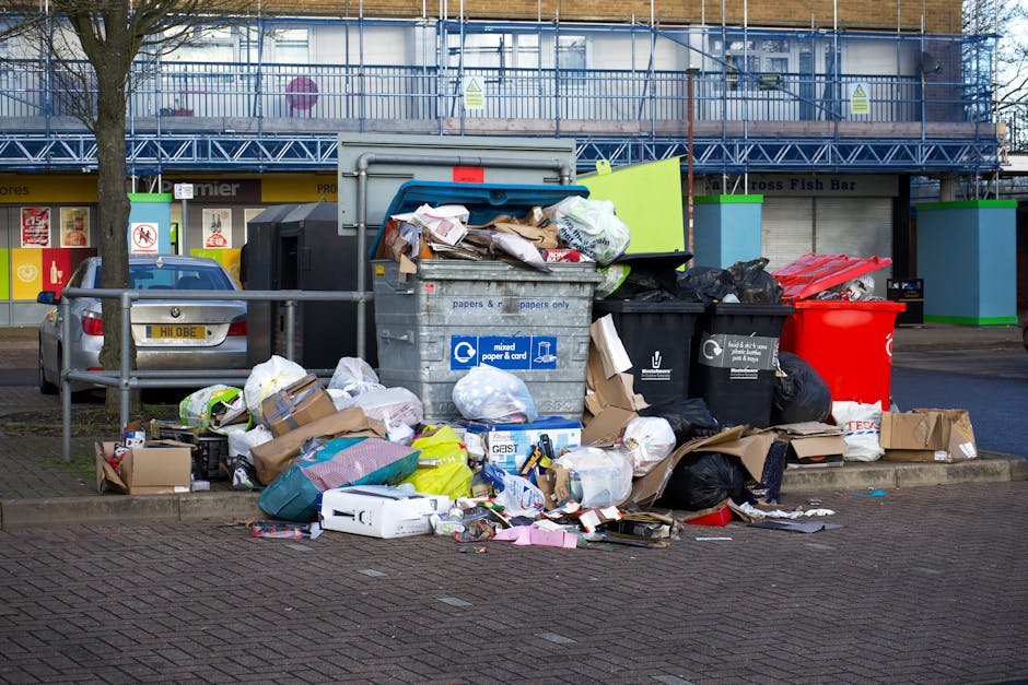 A collection of overflowing rubbish bins and scattered waste on a paved sidewalk area in an urban setting. The scene includes a large metal mixed paper and cardboard bin with its lid open, revealing crumpled paper, cardboard boxes, and plastic bags inside. Surrounding it are several black rubbish bags, some ripped open and spilling contents such as packaging, plastic bottles, and flattened cardboard. There is a red wheeled bin positioned to the right of the main containers. Additional loose refuse, including torn cardboard boxes, plastic wrapping, and paper spread across the pavement, suggests recent waste disposal activity. In the background, a low metal barrier separates the waste area from a parked grey car with a visible UK registration plate, and behind that, a building partially covered in scaffolding with blue netting. The scene is lit with natural daylight, emphasizing the cluttered and unmanaged appearance typical of an on-site waste collection area, relevant to private rubbish removal and alternative waste handling methods.