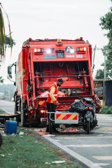 A waste disposal worker, dressed in a high-visibility yellow and red uniform, is standing at the rear of a large red rubbish collection truck parked on the side of a street. The worker appears to be operating or inspecting the compartment of the vehicle, which is open and shows internal mechanical components and hoppers. The scene includes a small black motorcycle parked beside the worker, with a black plastic bag placed on it, likely containing waste or materials to be collected. The street setting features a curb with a white painted line, some scattered debris and waste around the truck, and a grassy verge with a few trees in the background. The sky is overcast, casting diffuse natural light over the scene, which emphasizes the textures of the plastic bag, the metal surfaces of the truck, and the pavement. This image clearly depicts an operational waste collection process and may relate to private rubbish removal services provided by Waste Disposal Knightsbridge as part of alternative waste handling or on-site clearance.