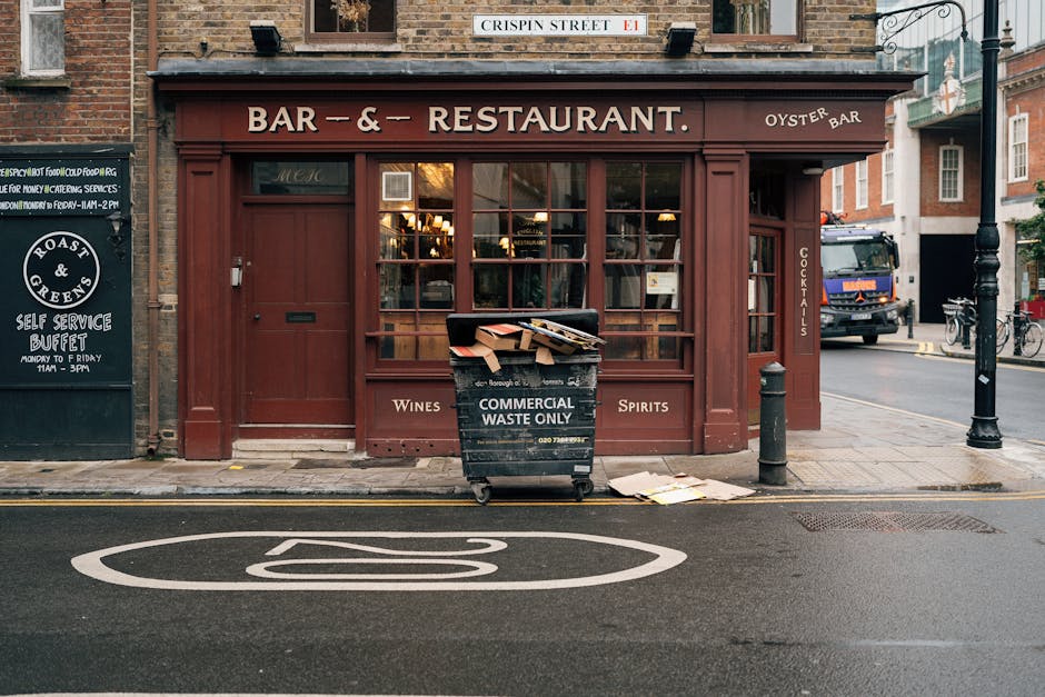 A waste disposal worker, dressed in a high-visibility yellow and red uniform, is standing at the rear of a large red rubbish collection truck parked on the side of a street. The worker appears to be operating or inspecting the compartment of the vehicle, which is open and shows internal mechanical components and hoppers. The scene includes a small black motorcycle parked beside the worker, with a black plastic bag placed on it, likely containing waste or materials to be collected. The street setting features a curb with a white painted line, some scattered debris and waste around the truck, and a grassy verge with a few trees in the background. The sky is overcast, casting diffuse natural light over the scene, which emphasizes the textures of the plastic bag, the metal surfaces of the truck, and the pavement. This image clearly depicts an operational waste collection process and may relate to private rubbish removal services provided by Waste Disposal Knightsbridge as part of alternative waste handling or on-site clearance.