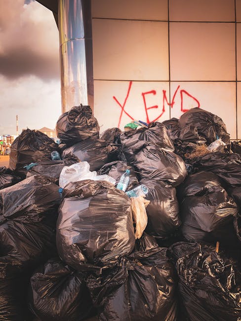 A large pile of black plastic rubbish bags is stacked against a modern building with a reflective metal surface and beige tiled exterior, some of the bags appear to be bulging and contain mixed waste. Several clear plastic water bottles are visible protruding from the top and sides of the bags, with some capped and others partially crushed. Behind the pile, red graffiti spelling 'XEND' is spray-painted on the tiled wall, and a cloudy sky with darkened clouds can be seen in the background. The scene suggests an unscheduled or improper waste disposal situation, potentially involving private or independent rubbish collection, which Waste Disposal Knightsbridge specialises in providing as an alternative to council services in the local area.