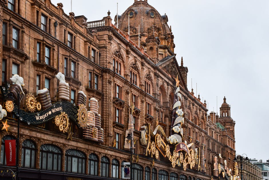 The image shows the detailed facade of a historic Victorian-style building constructed from reddish-brown sandstone, featuring ornate architectural elements including arched windows, decorative stone carvings, and small turrets on the roof. The upper section of the building is adorned with Christmas-themed decorations and lighted ornaments, with large, wrapped gift box props, golden stars, and illuminated gears contributing to a festive display. The scene is set during daytime with overcast, slightly dull weather, illuminating the intricate textures and finishes of the building’s stonework. The lower part of the building includes storefront windows, and in the background, the sky appears cloudy. The image subtly relates to waste removal or holiday event setups, reflecting how such decorations may require occasional private clearance or on-site removal services, as provided by Waste Disposal Knightsbridge, especially during peak festive seasons involving large-scale embellishments in urban settings.