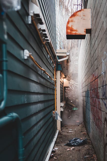 A narrow alleyway between a dark green, horizontally paneled building on the left and a taller, light-colored brick wall on the right. The alley is cluttered with small debris, including a black plastic bag, and is partially covered in dirt and grime. Along the green building, there are yellow and brown utility pipes and cables running horizontally, secured with brackets, with some sections showing oxidation. In the background, faint natural light filters through leafless tree branches, suggesting an outdoor setting during late autumn or winter. An outdoor light fixture or enclosure attached to the green building emits a soft glow. The scene captures a confined space often encountered in private waste disposal or off-site clearance jobs, relevant to independent rubbish removal services like those provided by Waste Disposal Knightsbridge, emphasizing the challenge of waste collection in restricted access areas.